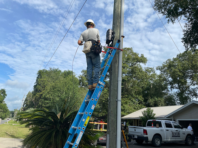 Electrician stands on a ladder up against an electric pole.
