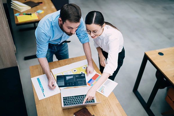 Two co-workers stand over a desk and work on their laptop in an office.