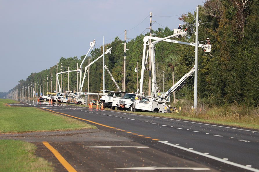 A fleet of boom trucks lift technicians to a row of electric poles.