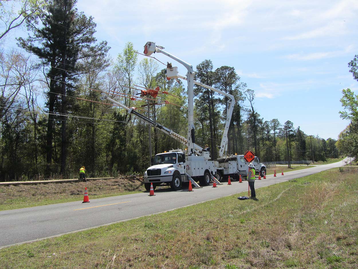 A electrical technician in a cherry picker lift working on a pole with other workers around.