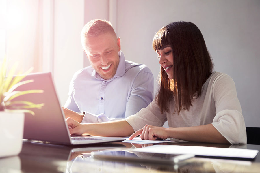 Two office workers sit at a desk using a laptop and pointing to papers.